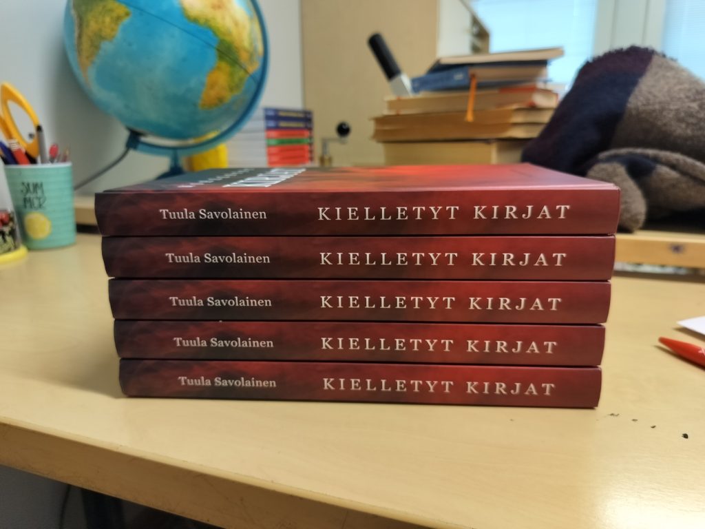 Vaalealla pöydällä pinottuna Kielletyt kirjat -teosta, kirjoista näkyy selkämys, taustalla pöydällä mm. karttapallo ja muita esineitä.