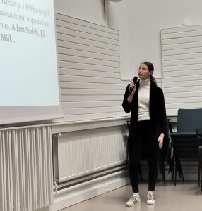 Elisa Viitasaari pitää esitystä Jokela-talon salissa. Hänellä on valkoinen poolopaita, musta bleiseri, mustat housut ja tennarit. Hän seisoo projektorin vieressä ja puhuu mikrofoniin katsoen valkokankaalle, jossa on tekstiä liittyen taloustieteeseen ja nimiä kuten Adam Smith ja J.S. Mill.