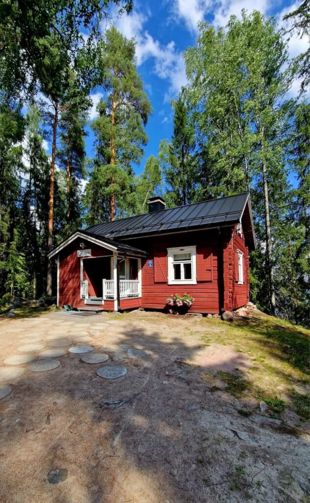 Punainen hirsimökki Kiviniemessä metsäisessä ympäristössä kirkkaan sinisen taivaan alla. Pihassa pyöreistä kivistä tehty polku, ja kuistin vieressä kukkaruukku täynnä kesäkukkia.