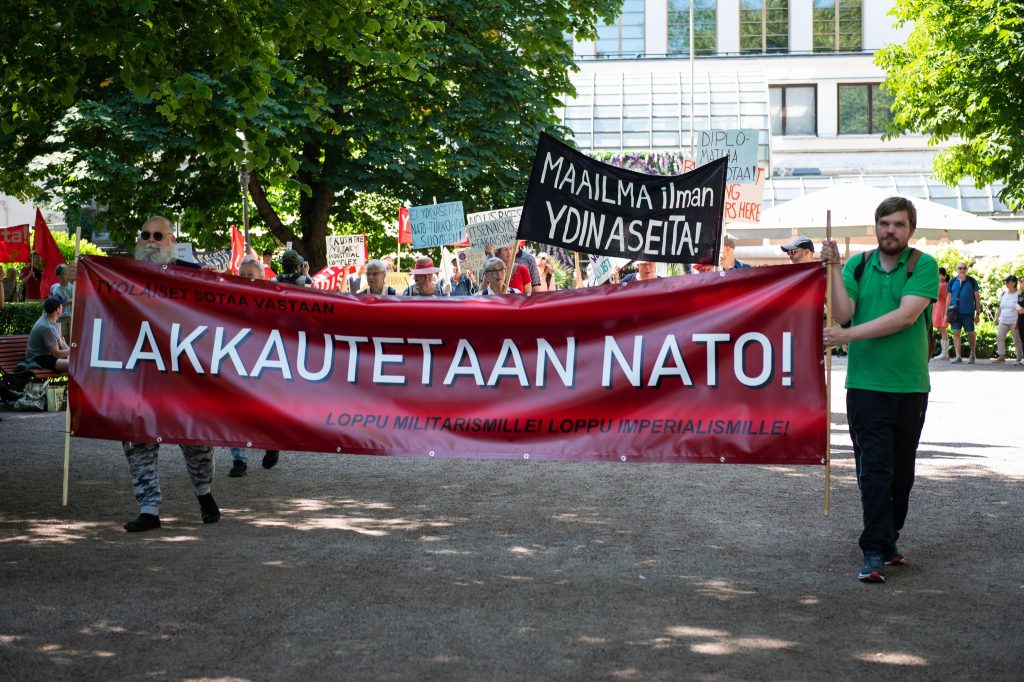 Mielenosoitus puistossa, jossa ihmiset kantavat suurta punaista banderollia tekstillä "Lakkautetaan NATO! Loppu militarismille! Loppu imperialismille!". Taustalla näkyy useita kylttejä, esimerkiksi "Maailma ilman ydinaseita!".