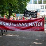 Mielenosoitus puistossa, jossa ihmiset kantavat suurta punaista banderollia tekstillä "Lakkautetaan NATO! Loppu militarismille! Loppu imperialismille!". Taustalla näkyy useita kylttejä, esimerkiksi "Maailma ilman ydinaseita!".