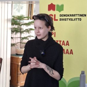 Bex Tapanainen alustamassa Marx-opistossa helmikuussa 2026, taustalla DSL:n roll-up.