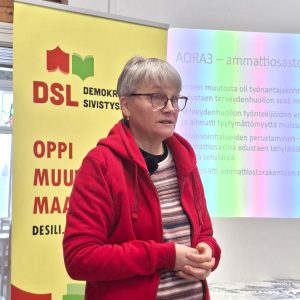 Mervi Grönfors alustamassa Marx-opistossa helmikuussa 2026, taustalla näkyy DSL:n roll-up.